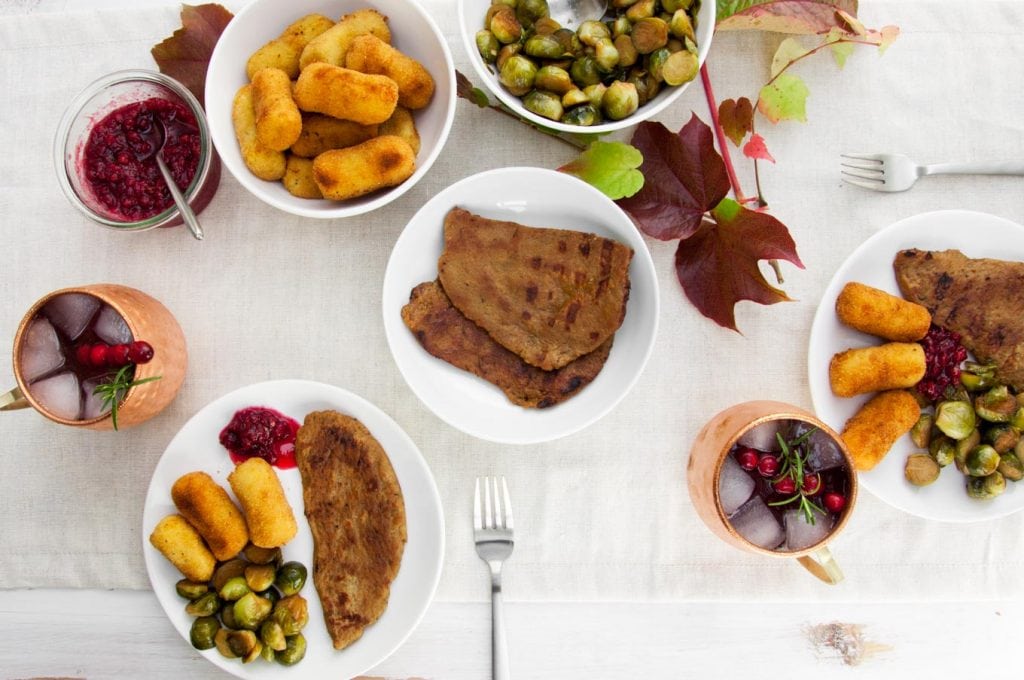 Vegan Thanksgiving Menu on a table with seitan steaks, brussels sprouts, potato croquettes, lingonberry jam and cranberry moscow mule