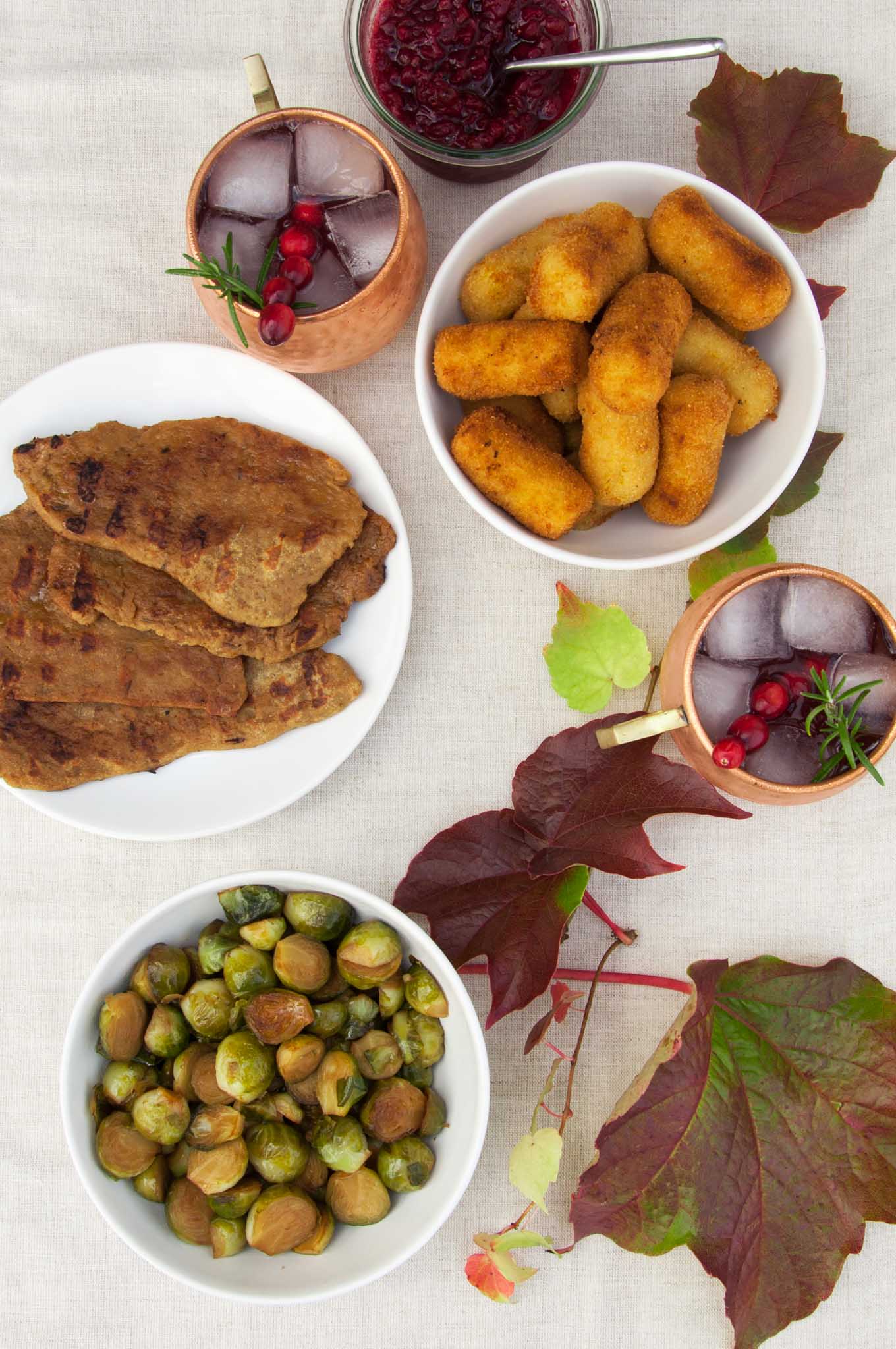 Vegan Thanksgiving Menu on a table with seitan steaks, brussels sprouts, potato croquettes, lingonberry jam and cranberry moscow mule