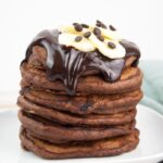 a stack of fluffy vegan chocolate banana pancakes topped with chocolate sauce, banana slices and chocolate chips