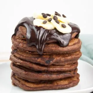 a stack of fluffy vegan chocolate banana pancakes topped with chocolate sauce, banana slices and chocolate chips