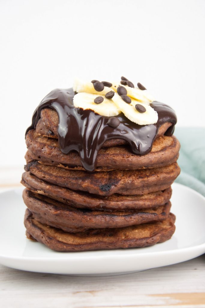 a stack of fluffy vegan chocolate banana pancakes topped with chocolate sauce, banana slices and chocolate chips