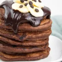 a stack of fluffy vegan chocolate banana pancakes topped with chocolate sauce, banana slices and chocolate chips