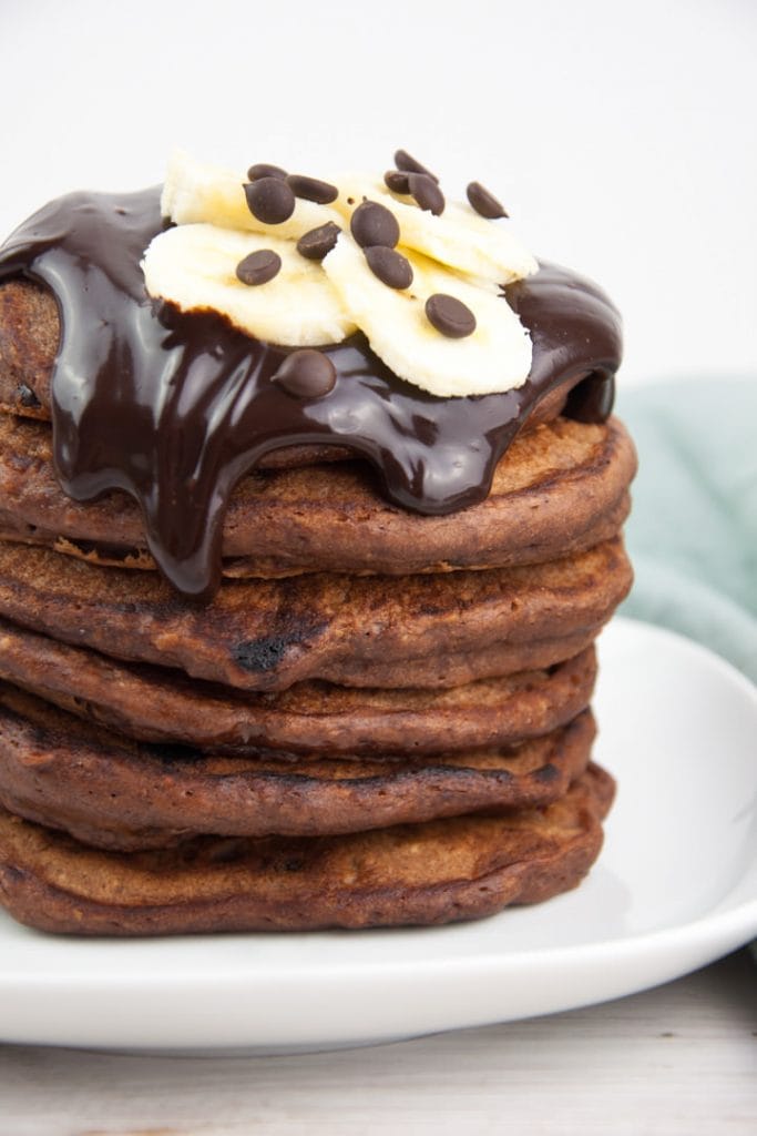 a stack of fluffy vegan chocolate banana pancakes topped with chocolate sauce, banana slices and chocolate chips